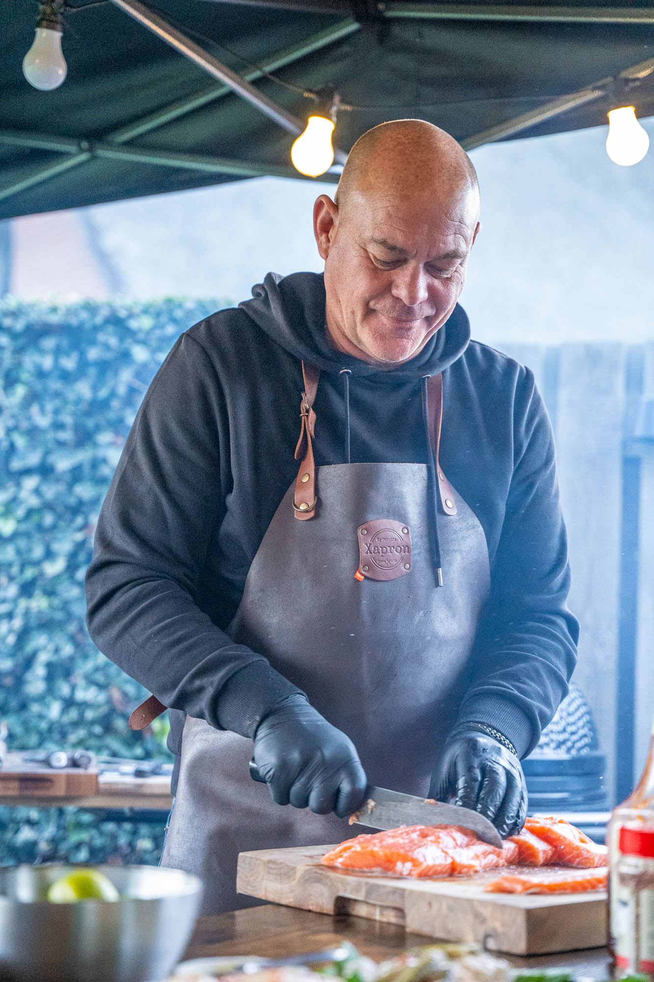 Michel van In Vuur en Vlam snijdt verse zalm op een houten plank, met zwarte handschoenen en leren schort